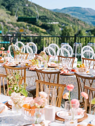 Outdoor tablescape at Viceroy Snowmass on the Lift Line Deck with two long tables, wooden chairs, florals, plates, and napkins in the foreground and a lift line in the background on a sunny day.